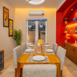 Celeste - Vio Stays - 3BHK Luxury Private Pool Villas In Baga - dining room with dining table and chairs, plates, fan & glasses. On the side - flower pots, paintings, wine glasses, tea cups, & air conditioner
